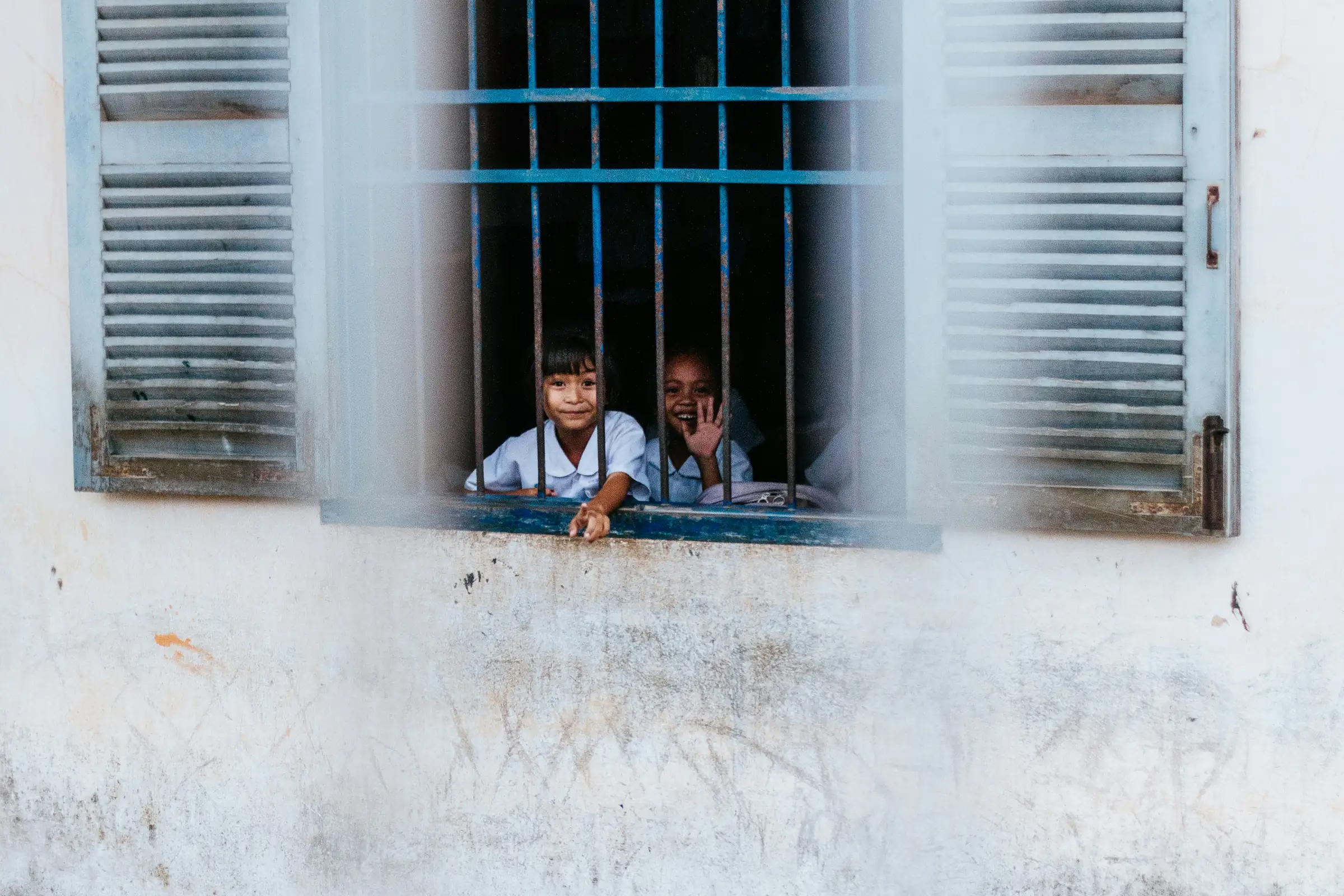 Two schoolchildren smiling and waving from a school window in Siem Reap, Cambodia