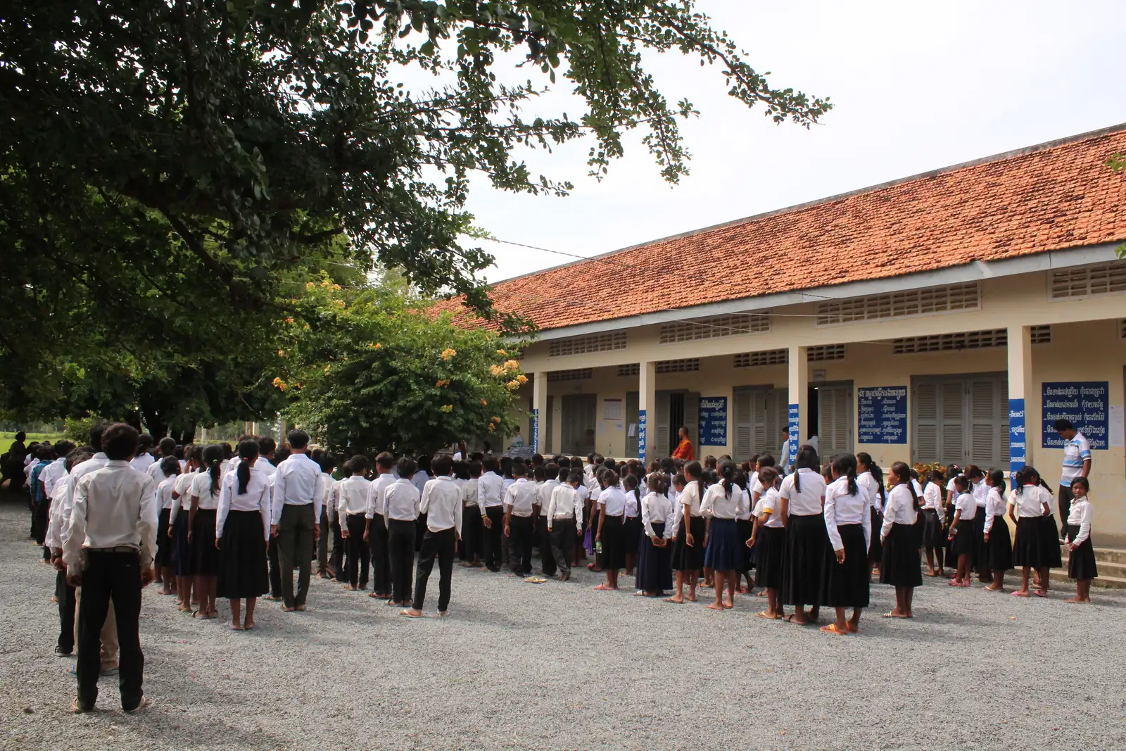 Students in white shirts lined up outside a red-roofed school building in Cambodia
